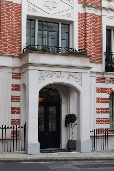 The image shows the front exterior of a red brick building with white architectural detailing, including a decorative archway framing a black door with a patterned glass panel. Above the door, there is a small white balcony with a wrought iron railing and a window behind it, partly obscured by a black-framed glass window with multiple panes. To the right of the entrance, a compact, round, pruned shrub in a dark gray pot is positioned near a black metal fence that runs along the sidewalk, which is clean and smooth. The building’s façade features intricate white decorative motifs with floral and scroll patterns, contrasting with the red brickwork. The lighting appears to be natural daylight, casting soft shadows and highlighting the textures of the brick and white embellishments. This setting suggests a well-maintained, elegant residential property, with subtle garden elements such as the potted shrub contributing to the overall curb appeal. Gardeners Mayfair specializes in garden maintenance and landscaping work near Berkeley Square, ensuring properties like this are kept meticulous and visually appealing.