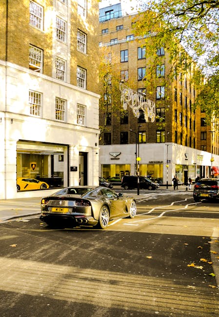 A city street scene in Mayfair with a focus on landscaping work visible in the foreground, featuring a well-maintained garden bed alongside the sidewalk filled with neatly trimmed green shrubs and small trees. The area has a clean, paved pathway bordered by a low hedge, with fallen autumn leaves scattered on the ground. Tall buildings with large windows line the street, casting warm sunlight that filters through the branches of nearby mature trees, giving a golden glow to the scene. Parked cars, including a yellow sports vehicle and a black sedan, are seen along the street, with a few pedestrians walking nearby. In the background, there's a brightly lit commercial shopfront, and decorative street lighting fixtures are visible hanging above the road. The overall environment reflects a combination of urban elegance and attentively maintained green spaces, showcasing professional gardening and landscaping attention by Gardeners Mayfair for outdoor aesthetic enhancement.