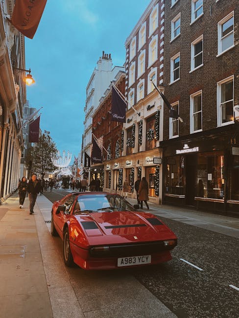 A red Ferrari sports car is parked on a busy city street in Mayfair, with pedestrians strolling along the pavement. The street is lined with multi-story buildings featuring large windows, some with decorative flags or banners hanging from their facades. The buildings display a mix of classic and modern architecture, with well-maintained facades and illuminated shopfronts. Trees are planted along the sidewalk, providing greenery amidst the urban environment. The sky above is partly cloudy, creating a soft daylight ambiance. Gardeners Mayfair's gardening and landscaping services focus on maintaining the aesthetics of such outdoor spaces, keeping lawns, pathways, and garden beds tidy and well-kept, which complements the upscale surroundings visible in this scene.